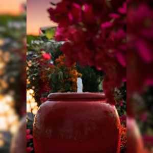 A vibrant red ceramic pot fountain bubbling water, surrounded by colorful flowers at sunset.