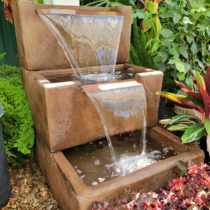 A two-tiered cascading water fountain with a modern, rectangular design surrounded by lush plants and greenery.