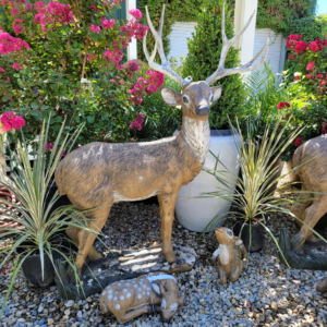 A life-sized deer statue with prominent antlers surrounded by smaller fawn statues, decorative plants, and blooming pink flowers.