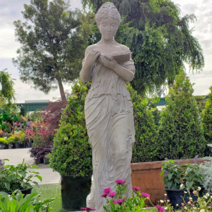 A classical stone statue of a woman reading a book, set amidst lush green plants and blooming flowers in an outdoor garden center.