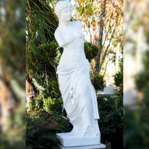 A white marble statue resembling Venus de Milo, set in a lush garden with greenery and natural light.