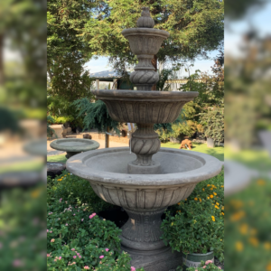 A three-tiered stone fountain surrounded by blooming flowers and lush greenery, set in a landscaped garden under a sunny sky.