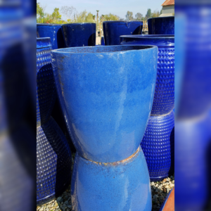 A tall, glossy blue ceramic garden pot with a smooth finish, surrounded by other blue pots in various sizes and textures, displayed outdoors on gravel.