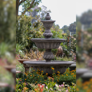A three-tiered stone fountain surrounded by blooming flowers and lush greenery in a garden setting.