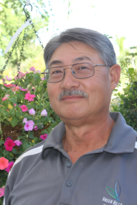 Leonard Ichimoto, Wholesale and Retail Sales representative at Green Hills Nursery, standing in front of colorful hanging flowers, wearing a gray Green Hills Nursery polo shirt.